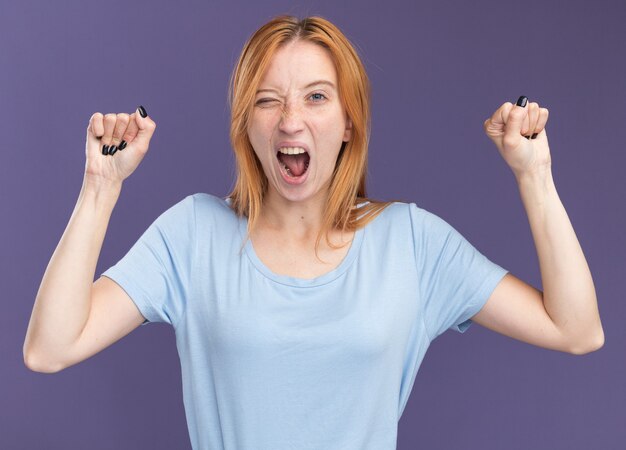 Annoyed young redhead ginger girl with freckles stands with raised fists isolated on purple wall with copy space