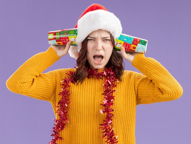 Free photo annoyed young slavic girl with santa hat and with garland around neck closing ears with paper cups isolated on purple background with copy space