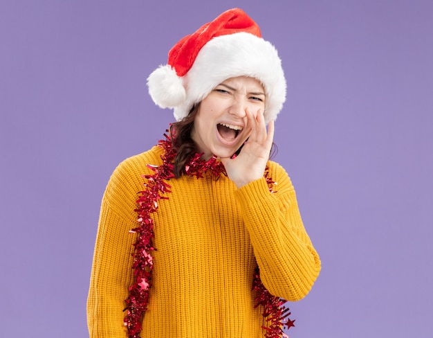 Annoyed young slavic girl with santa hat and with garland around neck holds hand close to mouth yelling at someone looking at camera isolated on purple background with copy space