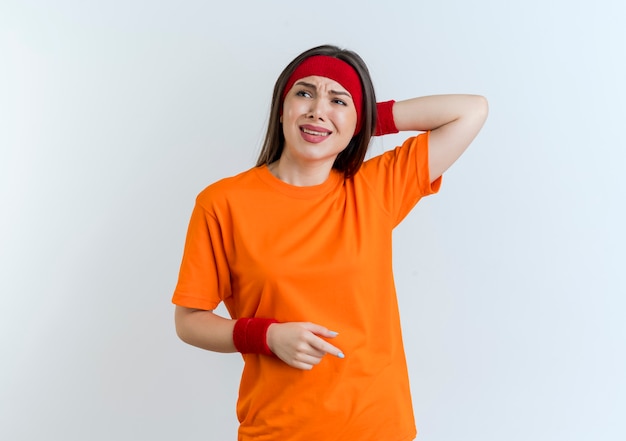 Annoyed young sporty woman wearing headband and wristbands keeping hand behind head looking at side isolated