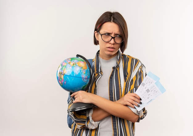 Бесплатное фото Раздраженная молодая студентка в очках и задней сумке с билетами на самолет и глобусом смотрит в сторону, изолированную на белом, с копией пространства