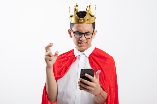 Annoyed young superhero boy in red cape wearing glasses and crown holding and looking at mobile phone keeping hand in air isolated on white background with copy space
