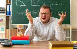 Free photo annoyed young teacher wearing glasses sitting at desk with school supplies in classroom keeping hands in air looking at front