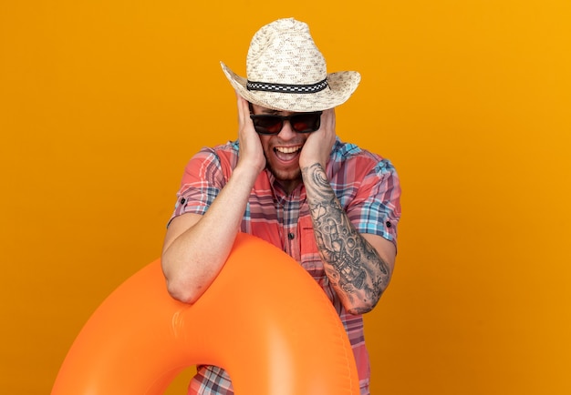Раздраженный молодой путешественник в соломенной пляжной шляпе в солнцезащитных очках держит кольцо для плавания и закрывает уши руками, изолированными на оранжевой стене с копией пространства