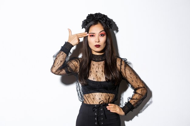 Annoyed young woman looking disappointed while making finger gun gesture over head, wearing halloween costume, standing over white background.