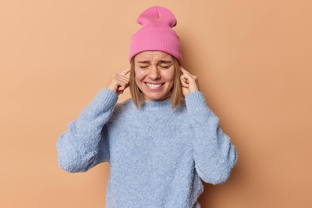 Annoyed young woman shuts ears looks distressed unwilling to hear noise stands displeased clenches teeth wears hat and jumper isolated over brown background Turn volume off Cannot hear anymore