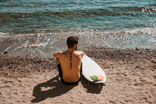 Anonymous man with surfboard sitting on sandy shore
