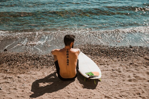 Anonymous man with surfboard sitting on sandy shore