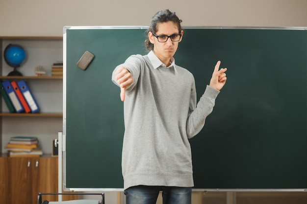 anpleased showing thumb up young male teacher standing on front of blackboard in classroom