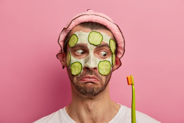 Anti aging procedure and teeth care concept. Close up shot of surprised unshaven European man wears cosmetic facial mask, looks shocked at toothbrush