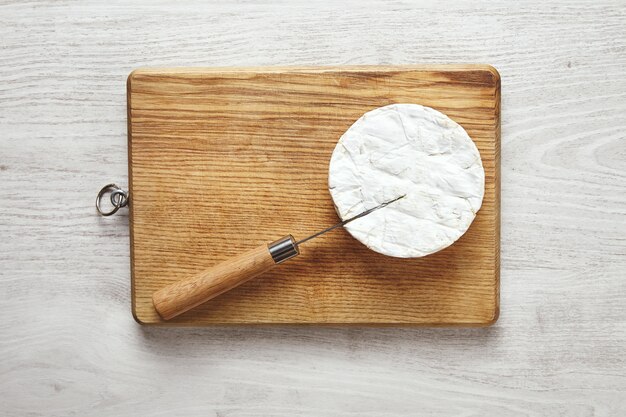 나무 손잡이가 달린 Antic 칼은 세 흰색 나무 테이블에 커팅 보드에 카망베르 치즈에 붙어 있습니다.