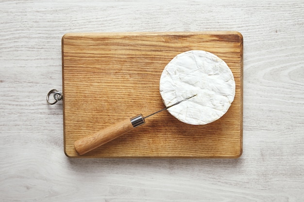 Antic knife with wooden handle stucks in camembert cheese on cutting board on aged white wooden table