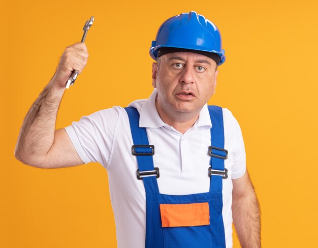 Anxious adult builder man in uniform holds wrench isolated on orange wall