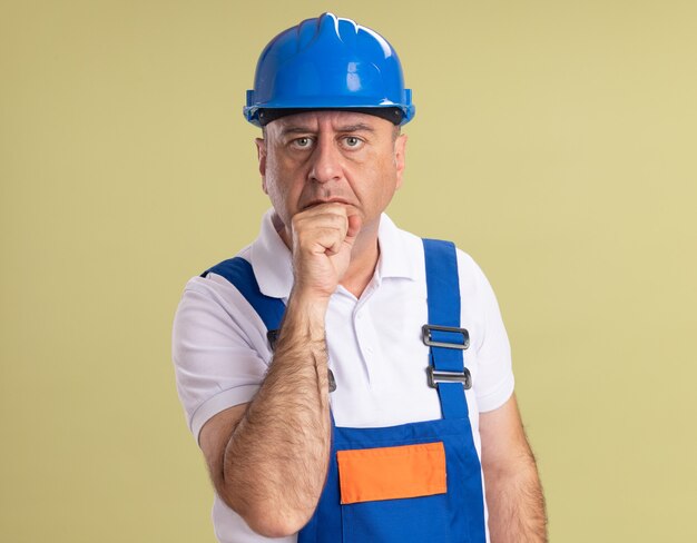 Anxious adult builder man in uniform puts fist on chin isolated on olive green wall