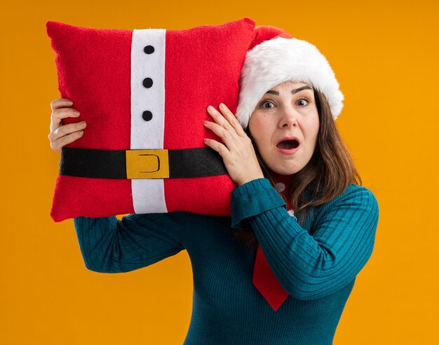 Anxious adult caucasian woman with santa hat and santa tie holding decorated pillow isolated on orange background with copy space