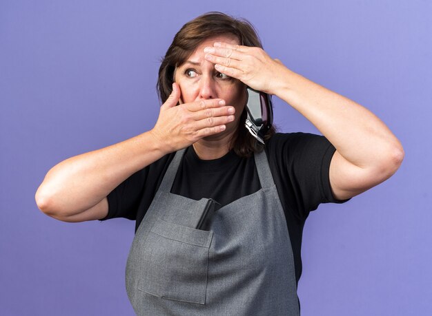 anxious adult female barber in uniform putting hand on forehead and on mouth holding hair clipper isolated on purple wall with copy space