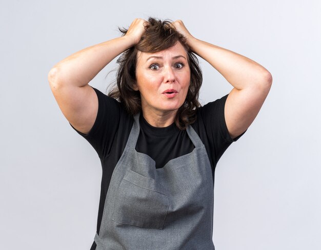 anxious adult female barber in uniform putting hands on head isolated on white wall with copy space