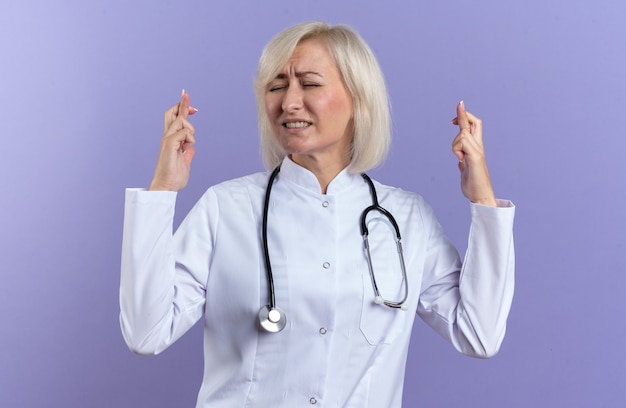 anxious adult female doctor in medical robe with stethoscope crossing fingers isolated on purple wall with copy space