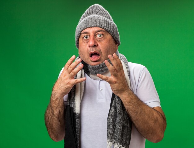 anxious adult ill caucasian man with scarf around neck wearing winter hat holding hands open isolated on green wall with copy space