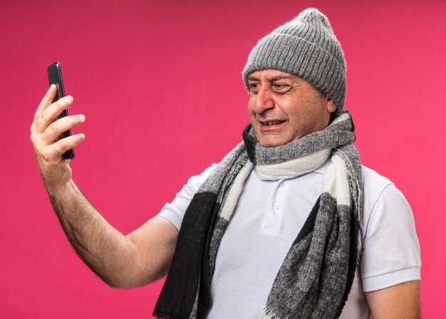 anxious adult ill caucasian man with scarf around neck wearing winter hat holding and looking at phone isolated on pink wall with copy space