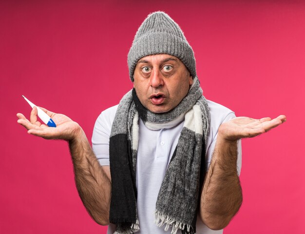 anxious adult ill caucasian man with scarf around neck wearing winter hat holding thermometer keeping hands open isolated on pink wall with copy space