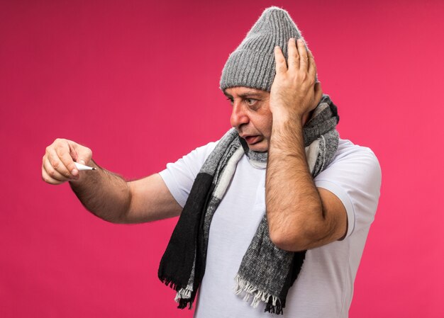 anxious adult ill caucasian man with scarf around neck wearing winter hat puts hand on head holding and looking at thermometer isolated on pink wall with copy space