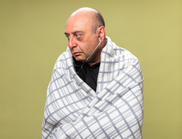 anxious adult ill caucasian man with stethoscope wrapped in plaid looking at side isolated on olive green wall with copy space