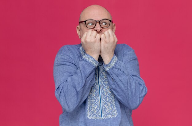 Anxious adult man in blue shirt wearing glasses biting his nails looking at side 