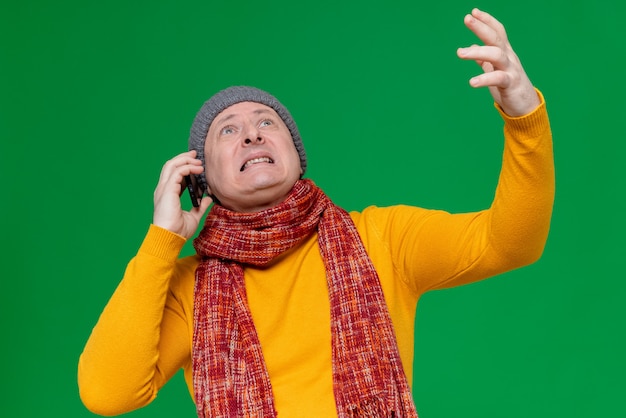 Uomo adulto ansioso con cappello invernale e sciarpa intorno al collo che parla al telefono guardando in alto