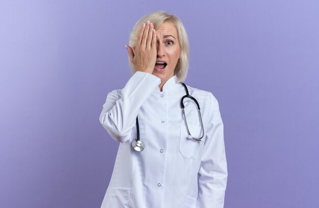 Anxious adult slavic female doctor in medical robe with stethoscope covering eye with hand isolated on purple background with copy space