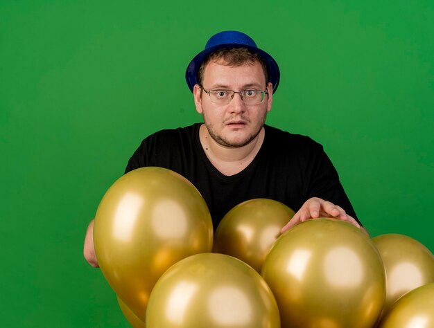 Anxious adult slavic man in optical glasses wearing blue party hat stands with helium balloons 