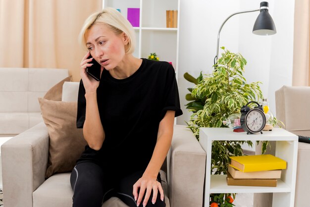 Anxious beautiful blonde russian woman sits on armchair talking on phone looking at side