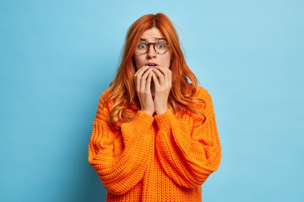 Anxious concerned disappointed redhead woman has terrible feared look holds breath as found out what happened hears bad horrible news dressed in knitted orange jumper.
