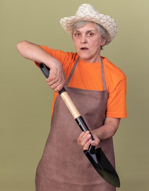 Anxious elderly female gardener wearing gardening hat holding spade on olive green