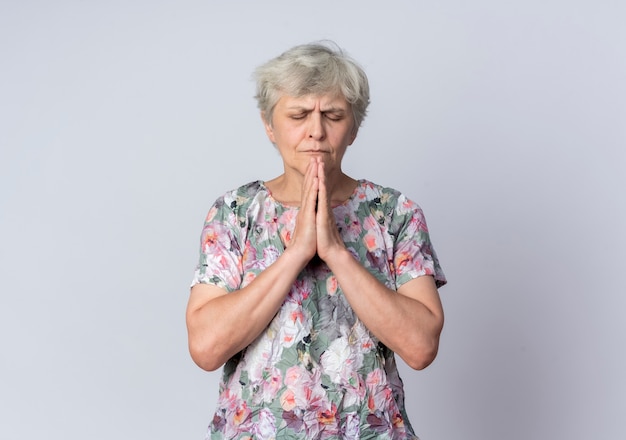 Anxious elderly woman holds hands together praying with closed eyes isolated on white wall