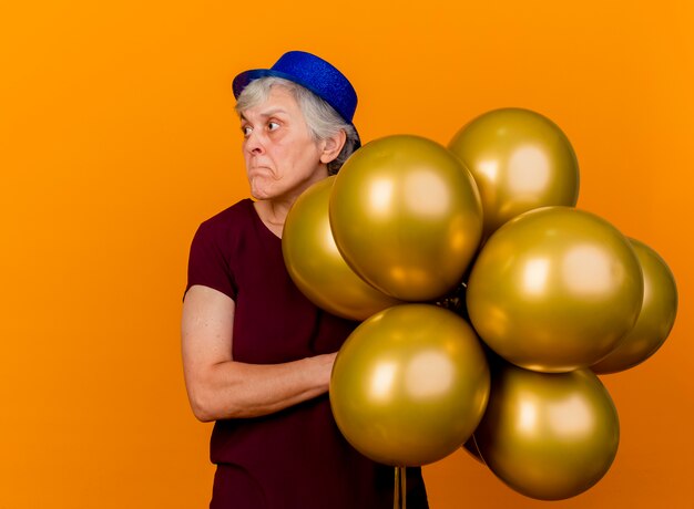 Anxious elderly woman wearing party hat holds helium balloons looking at side isolated on orange wall