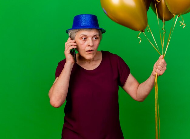 Anxious elderly woman wearing party hat holds helium balloons talking on phone on green