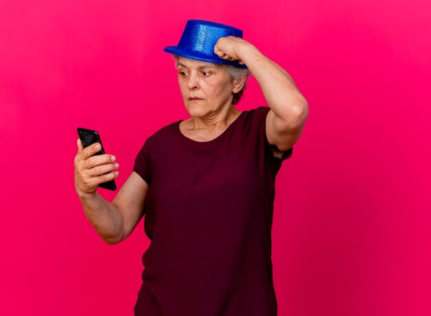 Anxious elderly woman wearing party hat puts fist on head looking at phone on pink