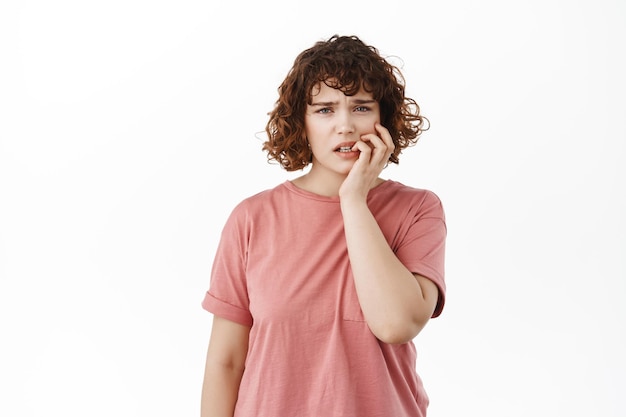 Anxious girl student biting fingernails and looking scared of consequences, fear of something, standing nervous and worried at camera, white background