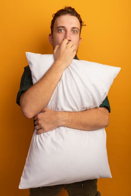 気になるハンサムなブロンドの男は、爪を噛み、オレンジ色の壁に分離された枕を保持します。