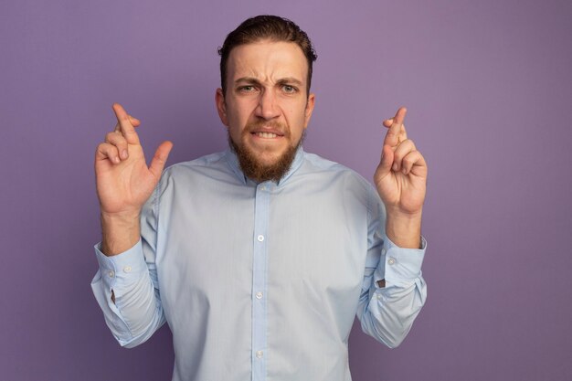 Anxious handsome blonde man crossing fingers isolated on purple wall