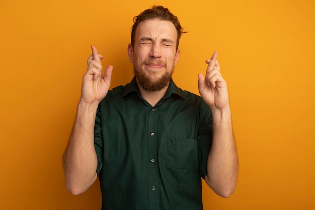 Anxious handsome blonde man stands with closed eyes crossing fingers isolated on orange wall