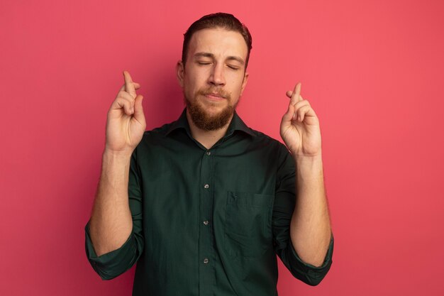 Free photo anxious handsome blonde man stands with crossed fingers isolated on pink wall