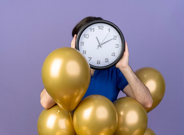 Anxious handsome caucasian man stands with helium balloons holding clock in front of face 