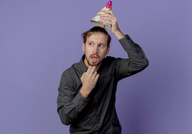 Anxious handsome man holds birthday cap over head looking at side isolated on purple wall