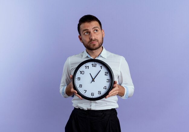 Anxious handsome man holds clock looking at side isolated on purple wall