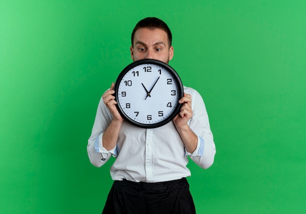 Anxious handsome man holds and looks at clock isolated on green wall