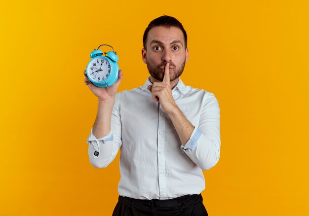 Anxious handsome man puts finger on mouth gesturing hush quiet holding alarm clock isolated on orange wall