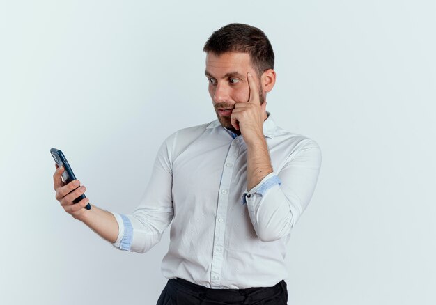 Anxious handsome man puts finger on temple holding and looking at phone isolated on white wall