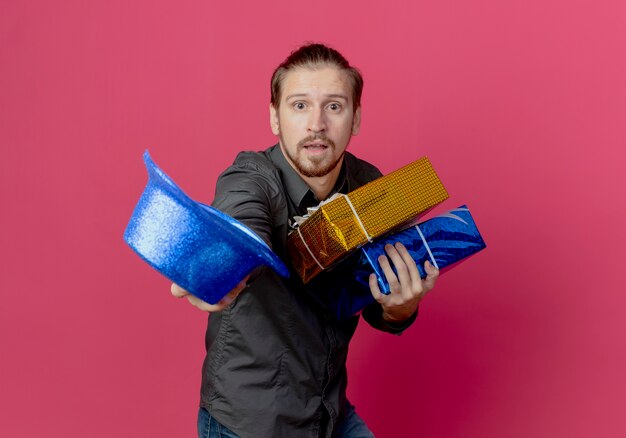 Anxious handsome man stands sideways holding gift boxes and blue hat isolated on pink wall